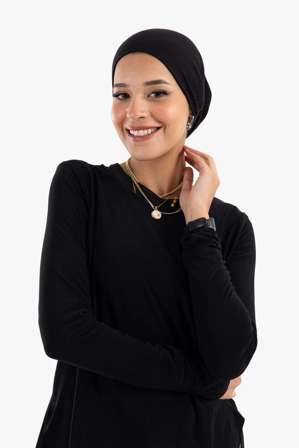 Woman wearing black long-sleeve shirt and black headscarf, smiling, accessorized with layered necklaces.