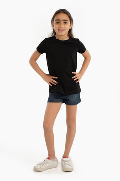 Girl wearing black short-sleeve top, denim shorts, and white sneakers on white background